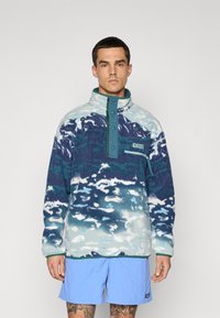 Mies, joka on pukeutunut kuvioituun sinivalkoiseen fleece-pulloveriin, jossa on napsautettavat napit, ja vaaleansinisiin shortseihin, seisoo tasaisen taustan edessä.