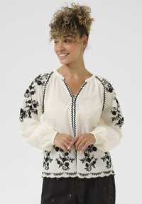 Hvid bluse med sort blomsterbroderi på ærmerne og kanten, der har en V-udskæring og rynkede manchetter, lavet af letvægtsstof.