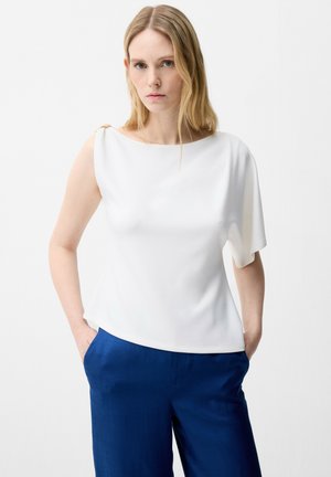 Kvinde med langt blond hår iført en hvid asymmetrisk kortærmet bluse og blå bukser, står med hænderne i lommerne mod en hvid baggrund.