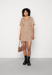 Vestido curto bege de manga curta com padrão ondulado castanho, com botões à frente e gola. Combinado com botas de combate escuras com atacadores e uma bolsa castanha clara.