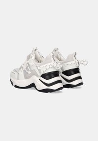 Zapatillas deportivas blancas y grises que presentan una mezcla de materiales, paneles de malla y una suela negra prominente con acentos en capas.