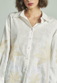 Camicia di lino bianca con motivi ricamati a foglia di palma beige. Collo classico, chiusura con bottoni e vestibilità rilassata con maniche lunghe.