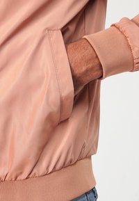 Jasnoróżowa kurtka bomberka o gładkiej fakturze, z ściągaczami na mankietach i boczną kieszenią, prezentująca minimalistyczny design i miękki materiał.