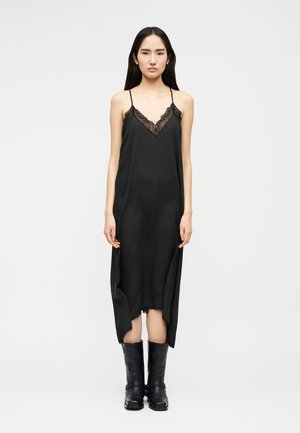 Mujer de pie, vestida con un vestido lencero negro sin mangas con ribete de encaje y botas de cuero negras hasta la mitad de la pantorrilla, sobre un fondo claro y sencillo.