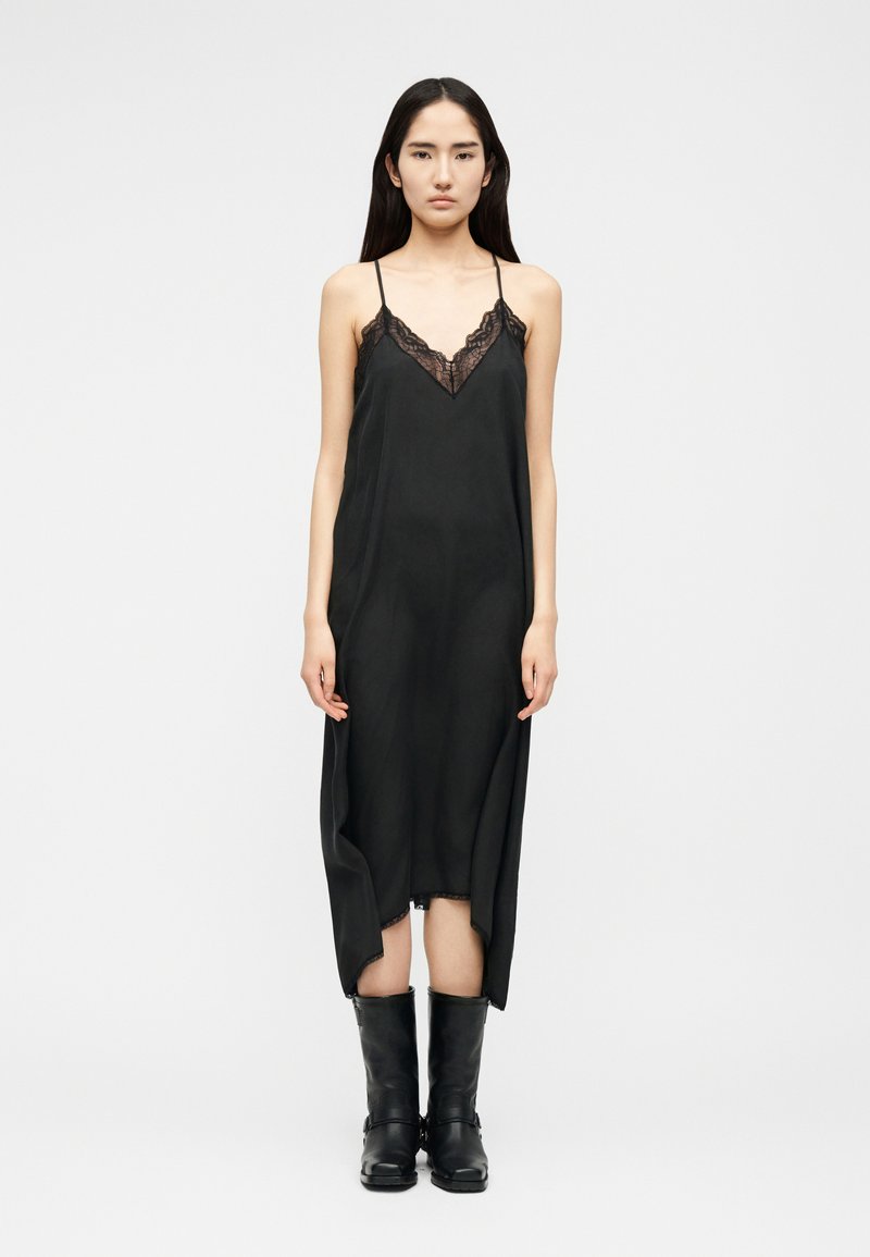 Γυναίκα που στέκεται φορώντας μαύρο αμάνικο φόρεμα με δαντέλα και μαύρες δερμάτινες μπότες μέχρι τη μέση της γάμπας, μπροστά σε απλό φωτεινό φόντο.