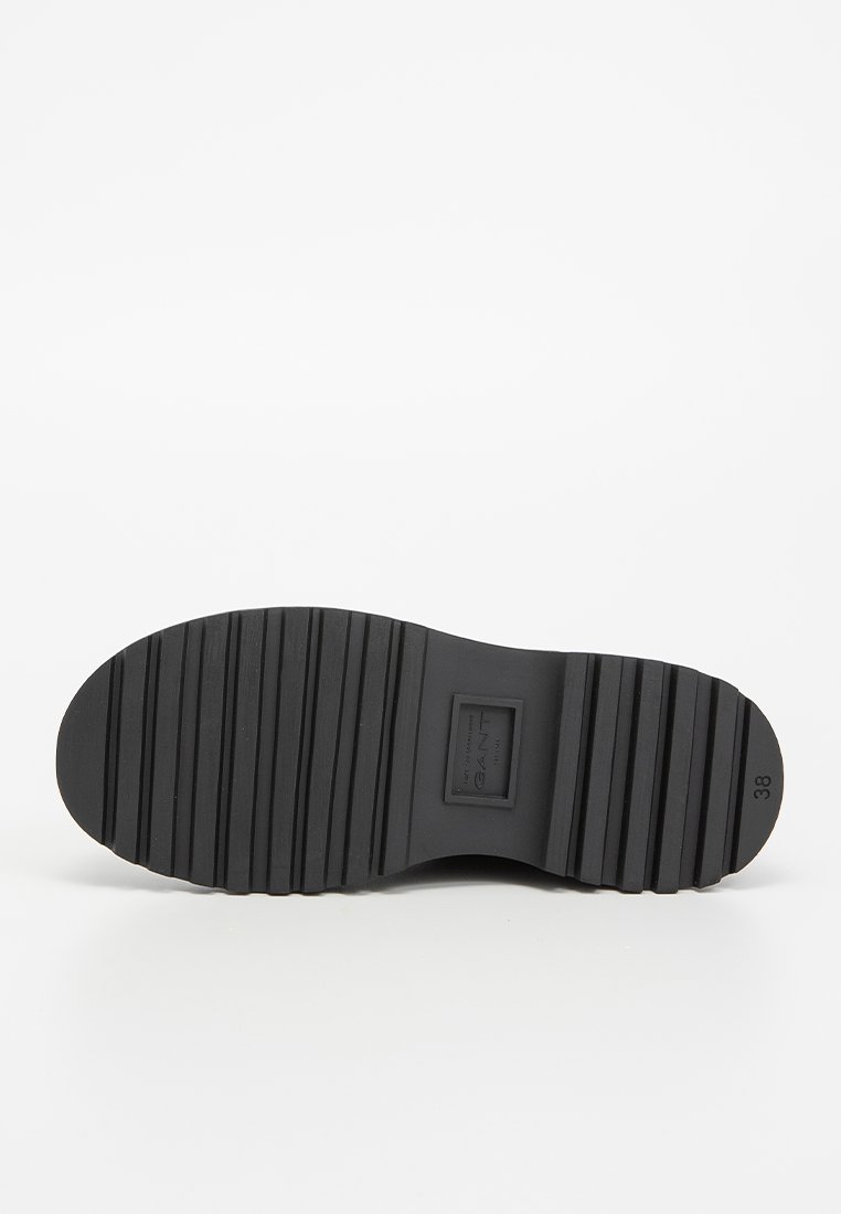 Suela de zapato de goma negra con un patrón texturizado y estriado, y un detalle circular en la punta. Tamaño marcado como 38. Logo de la marca visible en la suela.