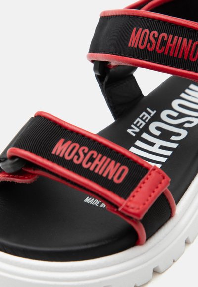 Μαύρο και κόκκινο πέδιλο Moschino για εφήβους με ρυθμιζόμενα λουράκια και λευκή χοντρή σόλα, με το λογότυπο Moschino στα λουράκια και στην εσωτερική σόλα.