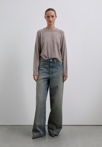 Vrouw die tegen een effen muur staat, gekleed in een lichtbruin gebreid sweater, wijde lichtblauwe jeans en puntige bruine schoenen.