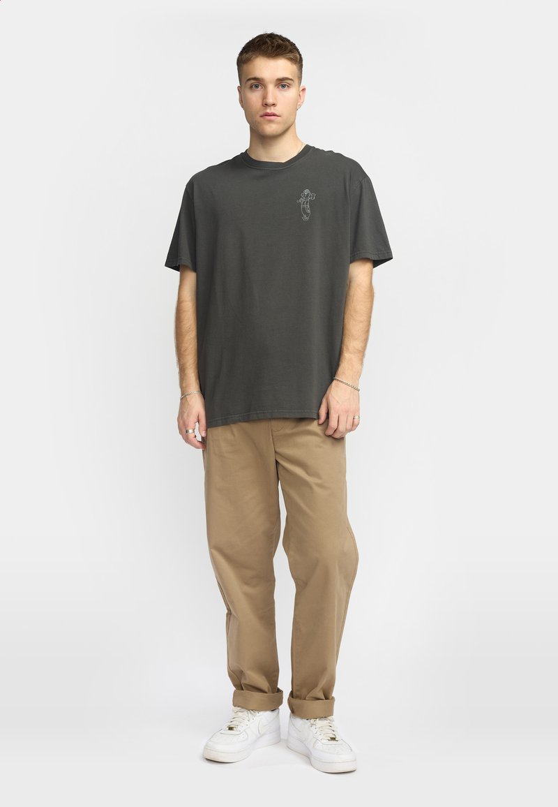 Camiseta de algodón gris oscuro con cuello redondo, que presenta un pequeño diseño gráfico blanco en el lado izquierdo del pecho, combinada con pantalones caquis claros y zapatillas blancas.