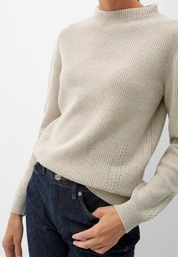 Beiger, gerippter Pullover mit hohem Kragen, verziert mit dekorativen Nähten an den Seiten und langen Ärmeln, kombiniert mit dunkler Jeans.