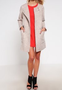 Cappotto esterno in misto lana beige chiaro con finitura testurizzata, chiusura doppia, tasche laterali e una vestibilità leggermente oversize. Vestito rosso sotto.