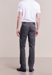 Man som bär mörkgrå byxor, vit t-shirt med kort ärm och svarta skor, står vänd bort från kameran framför en neutral bakgrund.
