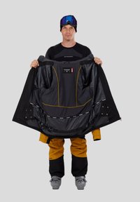 Zwart-gele skipully met een gladde binnenkant, getoond met uitgestrekte armen; heeft ritssluitingen, zakken en een mesh voering.