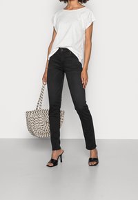 Camiseta blanca de manga corta, vaqueros negros ajustados, tacones abiertos negros y un bolso de mano tejido en blanco y negro, todo contra un fondo neutral.