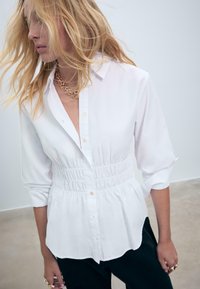 Donna bionda con capelli sciolti indossa una camicia bianca a maniche lunghe con bottoni e vita arricciata, insieme a collane in oro a strati.