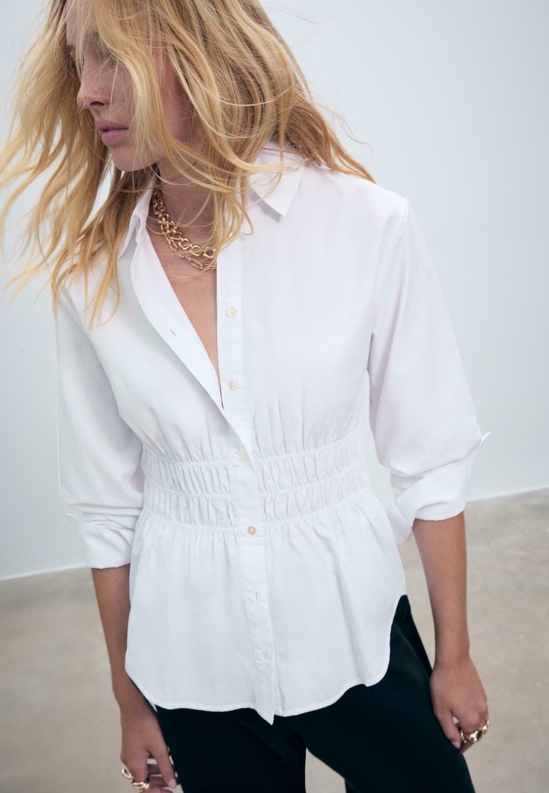 Donna bionda con capelli sciolti indossa una camicia bianca a maniche lunghe con bottoni e vita arricciata, insieme a collane in oro a strati.