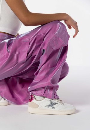 Pantaloni cargo lucidi viola con tasche e polsini elasticizzati. Abbinati a sneakers bianche con accenti argento e un logo bordeaux sul tallone.