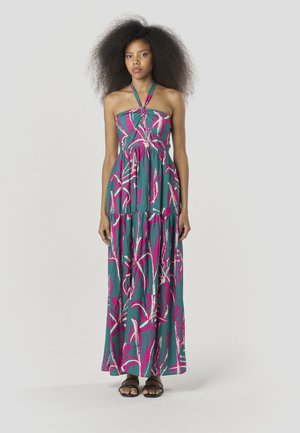 Maxi-jurk in teal met roze en witte abstracte patronen, halternekontwerp, gelaagde rok en lichtgewicht stof. Draagt zwarte sandalen.