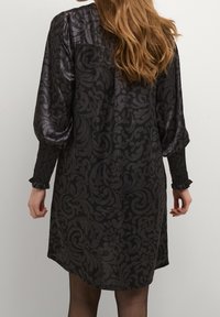 Černé šaty s lesklým povrchem, zdobené vzorovaným designem, nafouknutými rukávy a nařasenými manžetami. Délka po kolena s volným střihem.
