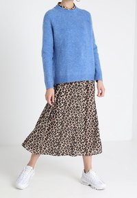 Kobieta w niebieskim swetrze, midi spódnicy w leoparda i białych trampkach stoi na tle białej ściany.