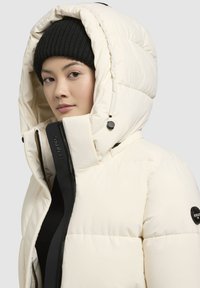 Vit puffjacka med stor huva, svart dragkedja och logotyp-patch. Tyget har en slät, glänsande yta och är isolerat för värme.