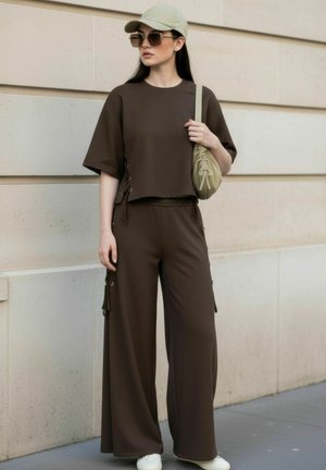 Femme en pantalon large marron et haut à manches courtes, portant une casquette beige, des lunettes de soleil oversize, des baskets blanches et tenant un sac à bandoulière beige.