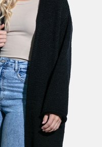 Czarna dziergana kardigan z otwartym przodem, długimi rękawami i teksturowanym materiałem. Zestawiony z beżową bluzką i niebieskimi dżinsami o wysokim stanie.