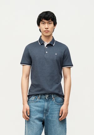 Giovane uomo che indossa una polo blu scuro con rifiniture bianche e jeans azzurri, in piedi contro uno sfondo chiaro e uniforme.