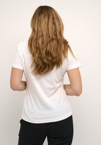 Valge lühikese varrukaga T-särk ümmarguse kaelusega, nähtud tagantpoolt. Kombineeritud mustade pükstega, millel on dekoratiivne õmblus taskutel.