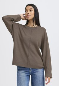 Pull en maille marron avec un col rond, des épaules tombantes et des poignets côtelés. Le matériau semble doux avec une coupe décontractée, associé à un jean bleu.