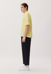 Camicetta gialla a maniche corte con motivi geometrici bianchi, abbinata a pantaloni blu navy e sneakers bianche. Tessuto liscio, vestibilità casual.