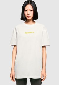 Weißes, übergroßes T-Shirt aus Baumwolle mit einem gelben "OPTIMIST"-Druck auf der Vorderseite. Kurze Ärmel und runder Halsausschnitt.