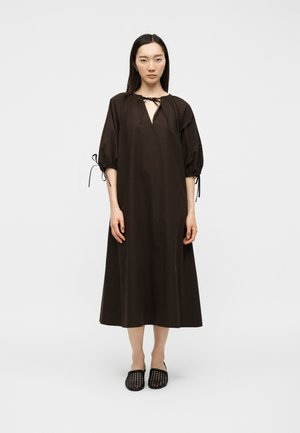 Kvinde står og bærer en løs mørkebrun midi kjole med halvt oppustede ærmer bundet ved manchetterne og sorte vævede slip-on sko.
