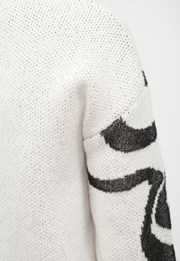 Baltas megztinis su juodu abstrakčiu raštu ant rankovės. Pagamintas iš tekstūruotos, laisvai austo audinio, turi atsipalaidavusią siluetą.