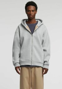 Grå hettejakke med glidelås, stor hette, oversized passform og ribbestrikkede mansjetter. Brukt over en marineblå t-skjorte, kombinert med beige bukser. Myk stofftekstur.