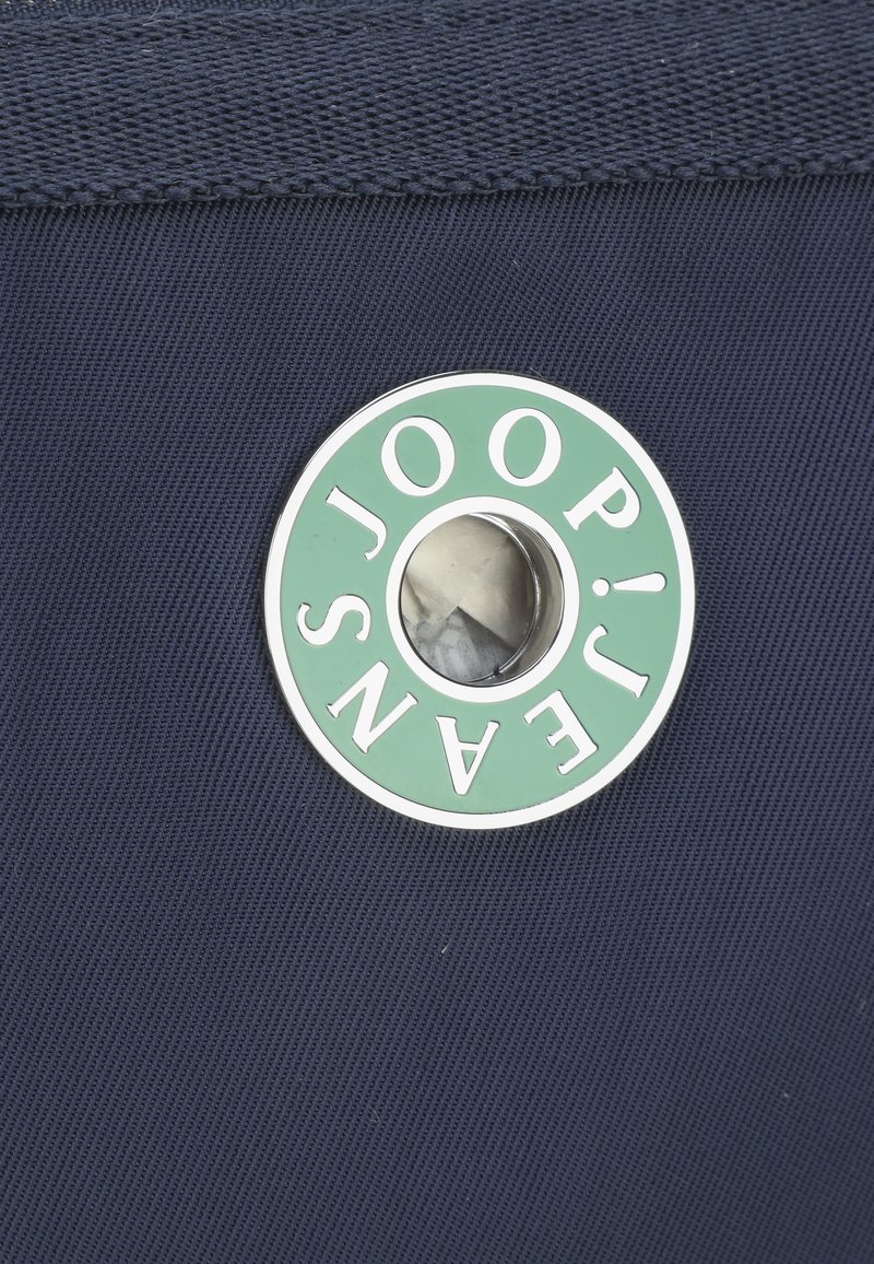 Navy-Stoff mit einem runden Metall-Logo, das grünen und weißen Text zeigt. Das Logo zeigt "JOOP!" und "JEANS" mit einem zentralen Loch-Detail.