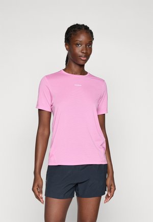 Femme se tenant devant un fond uni, portant un t-shirt de sport rose à manches courtes et un short noir, les cheveux tressés rabattus en arrière.