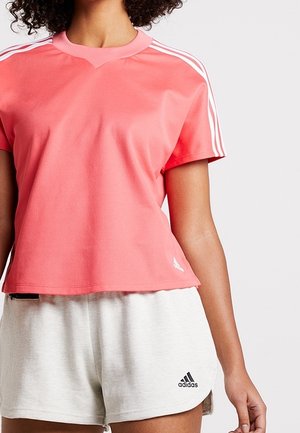 Femme portant un t-shirt Adidas corail à manches courtes avec des rayures blanches et un short Adidas gris clair.