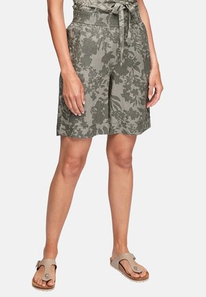 Lleva puestos unos pantalones cortos verdes con estampado floral, cintura anudada y sandalias beige sin talón, de pie contra un fondo liso.