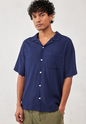 Giovane uomo con capelli ricci che indossa una camicia blu scuro a maniche corte con bottoni e pantaloni verde oliva, in piedi con una mano in tasca.