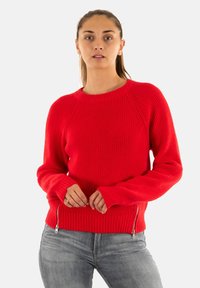 Donna che indossa un luminoso maglione rosso lavorato a maglia con cerniere in vita e jeans grigi, in piedi davanti a uno sfondo bianco semplice.