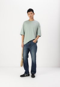 Lys grønn oversized T-skjorte, blå denimjeans, svarte sneakers med hvite detaljer, og en beige skulderveske. Enkel, uformell antrekk.