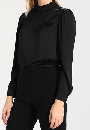 Blouse noire avec un col montant à volants, un empiècement structuré et des manches longues bouffantes. Tissu lisse avec un léger éclat, associé à un pantalon noir taille haute.