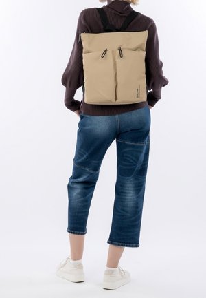 Sac à dos imperméable beige avec des fermetures à double zip, des bretelles réglables et une forme rectangulaire, porté sur le dos devant un fond neutre.