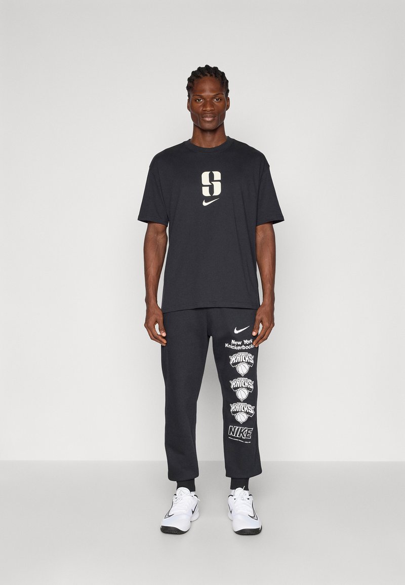 Schwarzes Baumwoll-T-Shirt mit großem weißen "S" und Nike-Logo. Passende dunkle Jogginghose mit weißen Knicks-Grafiken und Nike-Branding am Bein.