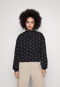 Geci scurt negru din material impermeabil, cu imprimeu alb pe toată suprafața, guler înalt, închidere cu fermoar și manșete elastice. Material neted și ușor.