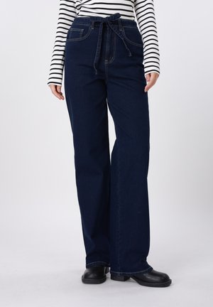 Dunkelblaue Jeans mit weitem Bein und gebundenem Taillengürtel, getragen mit schwarzen Schuhen und einem weißen Langarmshirt mit schwarzen horizontalen Streifen.