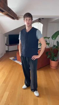 Colete matelassado azul-escuro com fecho de botões e dois bolsos, combinado com calças pretas largas e ténis brancos, situado numa sala com chão de madeira.