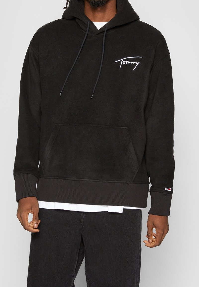 Svart fleece hoodie med en framficka, broderad "Tommy"-logotyp i vitt, dragsko på huvan och ribbstickade ärmslut. Buren över en vit skjorta.