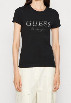 Femme portant un t-shirt noir ajusté avec le logo "GUESS Los Angeles" en lettres cursives pailletées, associé à un pantalon de couleur claire.